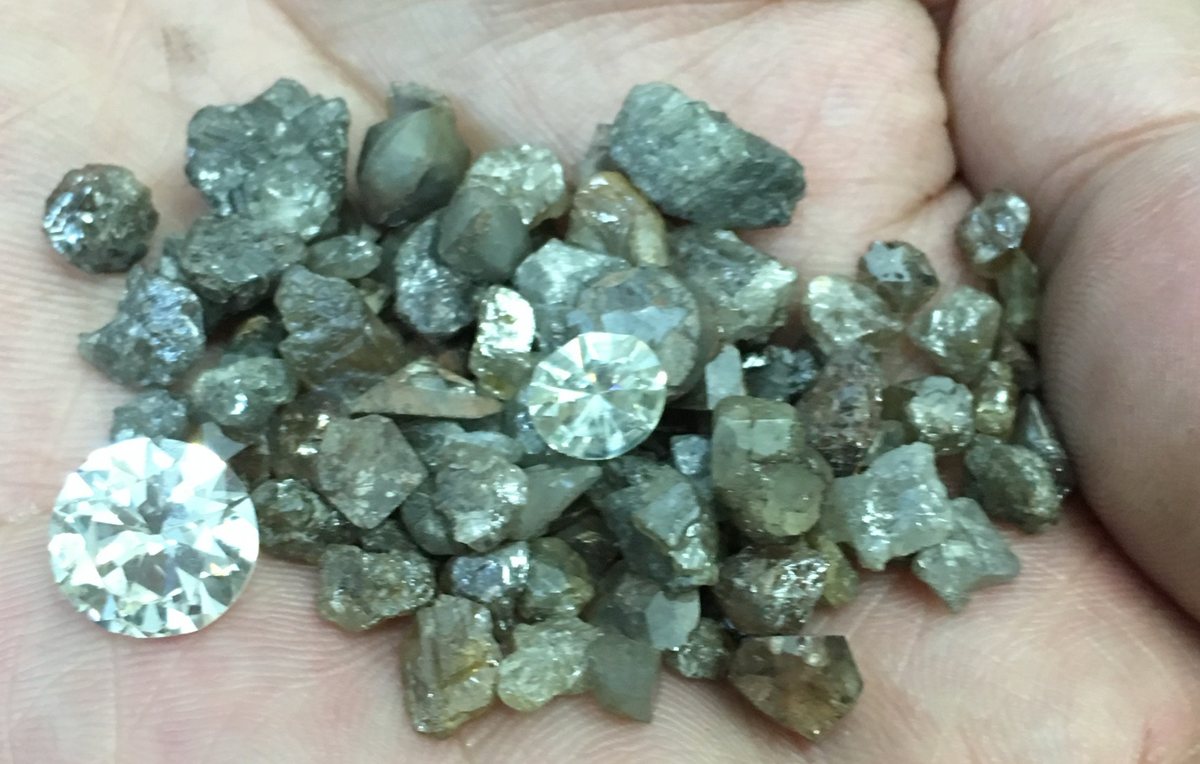 Grey and brown industrial-quality rough diamonds in a palm with two polished gem-quality round diamonds