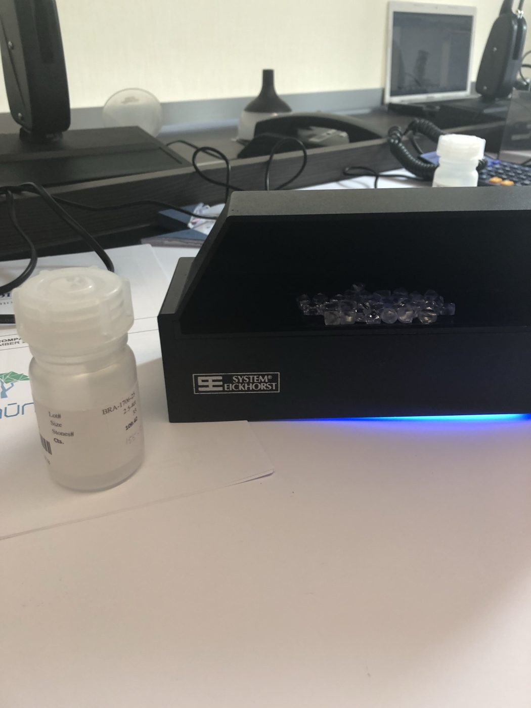 A parcel of rough diamonds inside a UV viewing box beside a labeled lot bottle